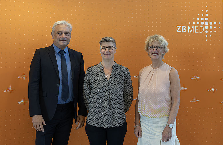 Gruppenbild vor orangener Wand mit Logo von ZB MED. Von links: Prof. Dr. Dietrich Rebholz-Schuhmann (Wiss. Direktor ZB MED), LMR'in Dr. Christiane Fricke (MKW), Prof. Dr. Juliane Fluck (Stellvertretende Direktorin komm. ZB MED)