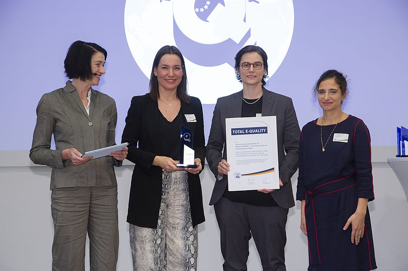 The photo shows four women standing next to each other: Dr Ulla Weber (Chair of the TOTAL E-QUALITY Executive Board), Christiane Hünten (Deputy Equal Opportunities Officer at ZB MED), Vanessa Scharf (Equal Opportunities Officer at ZB MED), Dr Susan Banihaschemi (Member of the TOTAL E-QUALITY Executive Board). Christiane Hünten holds a small statue, Vanessa Scharf a large certificate. The women stand in front of the TOTAL E-QUALITY logo.