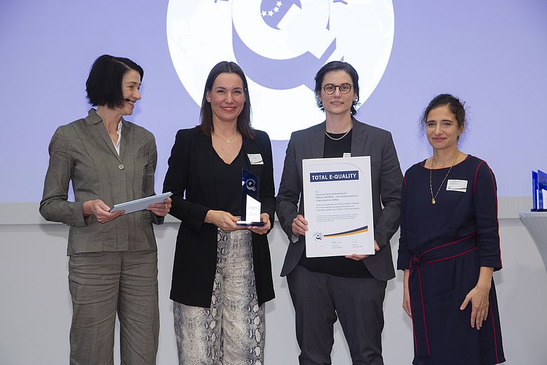 Auf dem Foto stehen vier Frauen nebeneinander: Dr. Ulla Weber (Vorstandsvorsitzende TOTAL E-QUALITY), Christiane Hünten (stellv. Gleichstellungsbeauftragte ZB MED), Vanessa Scharf (Gleichstellungsbeauftragte ZB MED), Dr. Susan Banihaschemi (Vorstand TOTAL E-QUALITY). Christiane Hünten hält eine kleine Statue in der Hand, Vanessa Scharf eine große Urkunde. Die Frauen stehen vor dem Logo von TOTAL E-QUALITY.